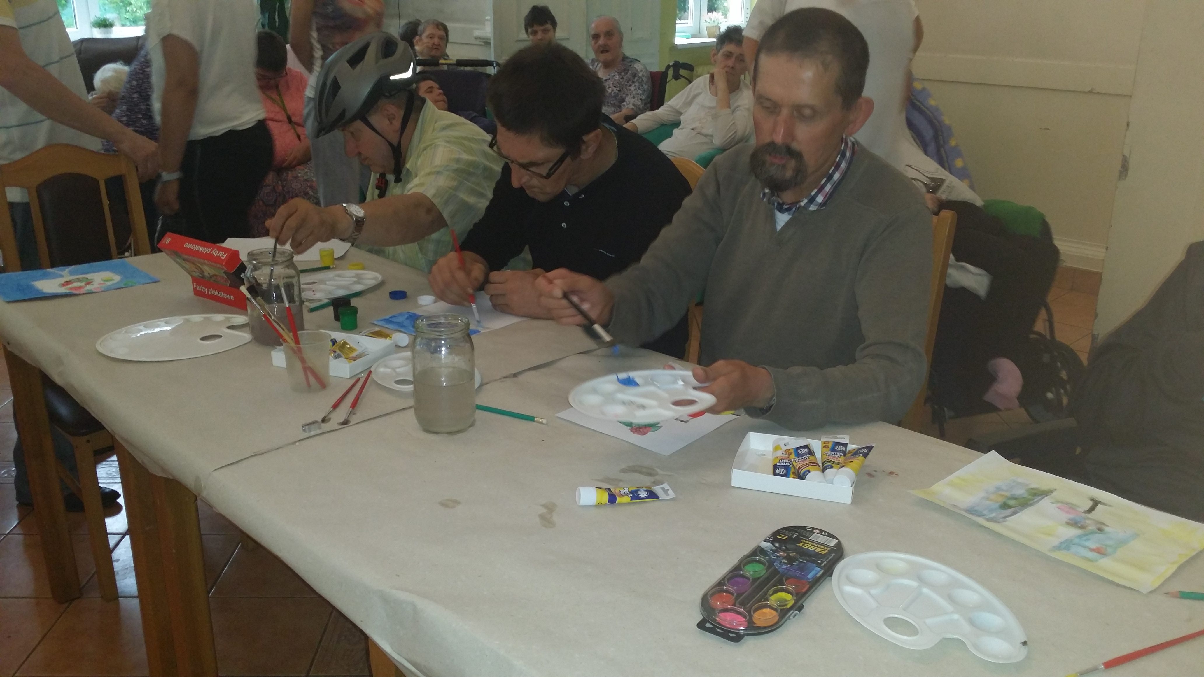 Uczestnicy pleneru malarskiego w Domu Pomocy Społecznej w Nowej Wsi siedząc przy stołach malują na kartce farbami kolorowe obrazy.