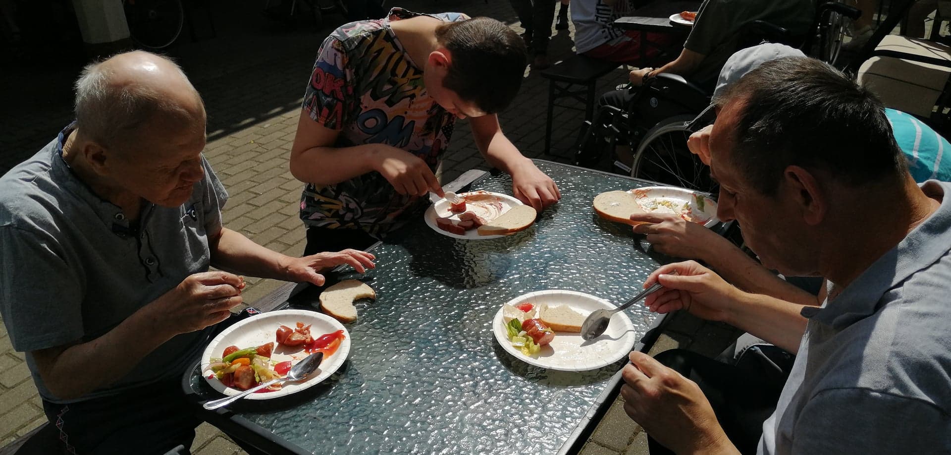 Mieszkańcy Domu Pomocy Społecznej w Chojnie siedzący przy stole jedzą kiełbasę z grilla.